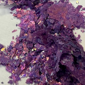 May include: Close-up of a pile of iridescent purple flakes. The flakes have a textured, layered appearance with hints of gold and pink. The material appears to be scattered on a light-colored surface.