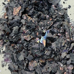 May include: Close-up of a pile of dark gray, irregularly shaped flakes. Some flakes have iridescent, colorful patches of purple, gold, and blue. The material appears to be a decorative element, possibly for crafts or art projects.