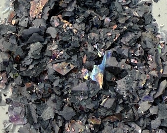 May include: Close-up of a pile of dark gray, irregularly shaped flakes. Some flakes have iridescent, colorful patches of purple, gold, and blue. The material appears to be a decorative element, possibly for crafts or art projects.