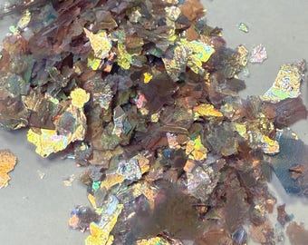 May include: Iridescent flakes in shades of gold, bronze, and silver spill from a clear bag. The flakes have a textured, fractured appearance, catching the light and creating a shimmering effect. The material appears to be a decorative craft supply.