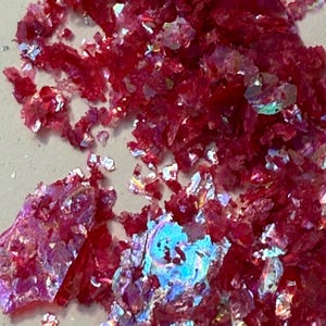May include: Close-up of scattered, iridescent red flakes. The flakes have a crystalline appearance with a reflective, shimmering quality. The fragments vary in size and shape, creating a textured, scattered pattern.