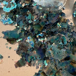 May include: Close-up of iridescent, teal-colored flakes, possibly for crafting or decorative purposes. The flakes have a fractured, irregular shape and reflect light, creating a shimmering effect. Some flakes are contained in a clear plastic container.