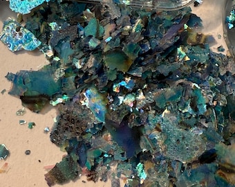 Puede incluir: Primer plano de escamas iridiscentes de color verde azulado, posiblemente para manualidades o fines decorativos. Las escamas tienen una forma fracturada e irregular y reflejan la luz, creando un efecto brillante. Algunas escamas están contenidas en un recipiente de plástico transparente.