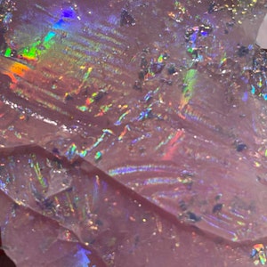 May include: A close-up of a piece of iridescent purple opal with a rainbow sheen. The opal has a rough, textured surface.