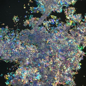 May include: Iridescent, shimmering flakes scattered on a black surface. The flakes reflect a spectrum of colors, including blues, greens, and purples, creating a dazzling effect. The material appears to be thin and delicate.