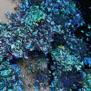 May include: Close-up of iridescent blue and green glitter flakes scattered on a white surface. The glitter has a metallic sheen and reflects light.