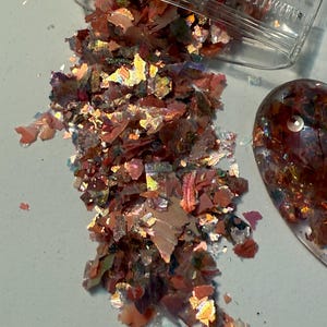 May include: A close-up of iridescent flakes in shades of red, orange, and gold, spilling from a clear plastic container. A small, oval-shaped object with similar flakes is visible to the right. The flakes have a reflective, shimmering quality.