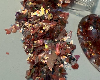 Puede incluir: Primer plano de escamas iridiscentes en tonos rojos, naranjas y dorados, derramándose de un recipiente de plástico transparente. Un pequeño objeto ovalado con escamas similares es visible. Las escamas tienen una cualidad reflectante y brillante.