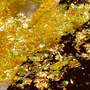 May include: Close-up of iridescent gold glitter flakes, some of which are scattered on a black surface.