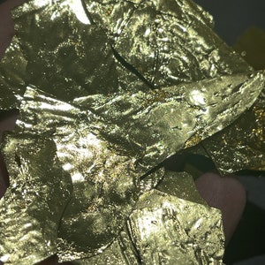 May include: A pile of gold leaf flakes, with a textured surface, in a hand.