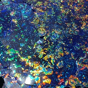May include: Close-up of iridescent blue, green, and gold glitter flakes on a black background. The glitter is scattered randomly and creates a shimmering effect.