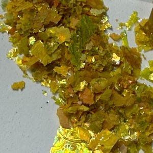 May include: Close-up of scattered, iridescent gold and green flakes. The flakes have a textured, irregular shape and reflect light, creating a shimmering effect. The flakes are scattered on a light gray surface.