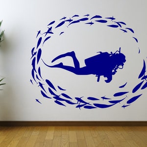 May include: A blue wall sticker depicting a scuba diver encircled by a shoal of fish. The diver is centrally positioned, with the fish arranged in a circular formation. The design is on a white wall, with a potted plant and wooden flooring.