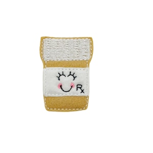 May include: A yellow felt pill bottle with a white label and a smiling face with pink cheeks. The label also has the letter "R" on it.