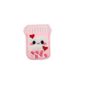 May include: A pink felt patch in the shape of a pill bottle with a smiling face and red heart accents. The bottle contains red and white striped candies. The patch is designed with embroidered details and a white background.