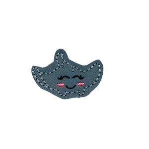 May include: A gray felt patch with a smiling manta ray design. The manta ray has pink cheeks and black eyes.