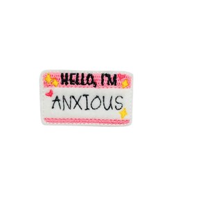 May include: Pink and white embroidered name tag with the text "Hello, I'm Anxious" in black lettering. The tag has yellow stars and pink hearts.