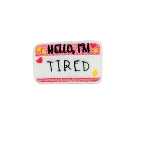 May include: White felt name tag with pink trim and embroidered text that reads "Hello, I'm Tired". The tag has a red heart and yellow stars.