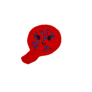 May include: A red felt applique in the shape of a stylized face with a handle. The face features blue embroidered details resembling branches and black embroidered eyes. The applique is a whimsical, handmade item.