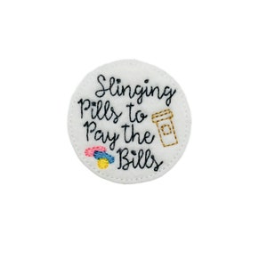 May include: White felt circle with black embroidered text that reads "Slinging Pills to Pay the Bills". A small image of a coffee cup and three pills are also embroidered on the circle.