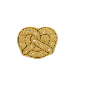 May include: A brown felt pretzel with white embroidery, resembling a pretzel with sprinkles.