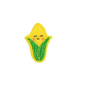 May include: A yellow felt corn cob with a green felt husk and a smiling face. The corn cob has pink cheeks and black eyes.