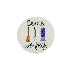 May include: A white circle with a glitter texture and a black embroidered design. The design includes three brooms, a vacuum cleaner, and the text "Come, we fly!"