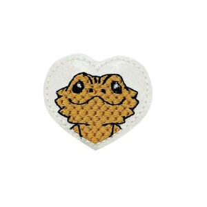 May include: A white heart-shaped patch with a brown bearded dragon embroidered on it. The dragon has large eyes and a friendly expression.