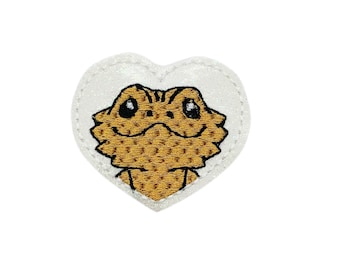 May include: A white heart-shaped patch with a brown bearded dragon embroidered on it. The dragon has large eyes and a friendly expression.