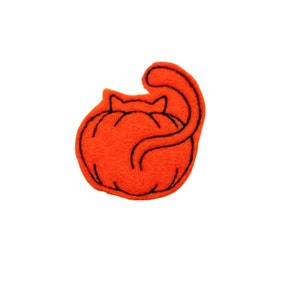 May include: Orange felt cat curled up in a ball, with black embroidered details. The cat is shaped like a pumpkin.