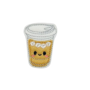 May include: A white and brown felt coffee cup with a smiling face and a white swirl on top. The cup is embroidered with a silver thread.