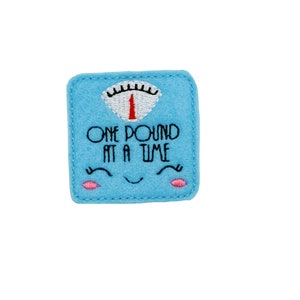 May include: A light blue felt square with embroidered text and a smiling face. The text reads "ONE POUND AT A TIME" above the smiling face. The top of the square features a scale design with the number 1.