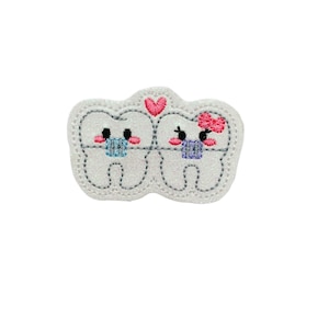 May include: A white felt patch with two cartoon teeth wearing braces and a pink heart above them. The teeth have pink cheeks and black eyes. The female tooth has a pink bow.