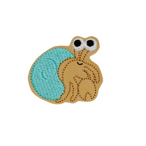 May include: A whimsical embroidered patch featuring a cartoon hermit crab. The crab has a tan body with a light blue shell and large, googly eyes. The design is outlined with brown stitching, creating a playful and detailed image.
