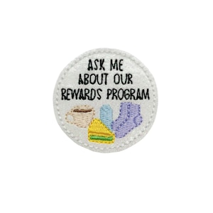 May include: White round button with black text that reads "Ask me about our rewards program". The button has a white background with a glittery texture. There are three embroidered images on the button: a cup of coffee, a pair of socks, and a slice of cake.