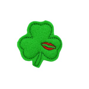 May include: A green felt shamrock with a red embroidered kiss on one of the leaves.
