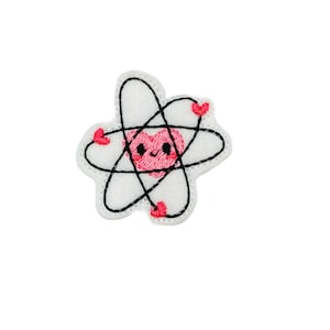 May include: A white felt patch with an embroidered black atom symbol. The atom has a pink heart in the center with a smiling face. There are four pink hearts around the atom.