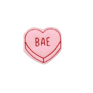 May include: Pink felt heart-shaped candy with red embroidery outlining the heart and the word "BAE" inside.