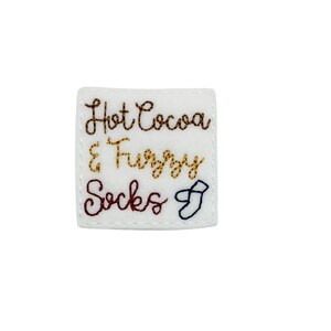 May include: White felt patch with embroidered text that reads "Hot Cocoa & Fuzzy Socks" with a small embroidered sock icon.