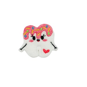 May include: A white felt tooth with pink frosting and colorful sprinkles. The tooth has a happy face with eyelashes and a red heart on its chest.