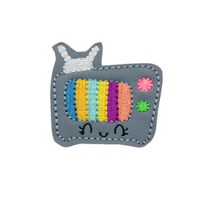 May include: A gray felt embroidered patch of a cartoon television with a rainbow screen, white bunny rabbit antenna, and a smiling face.