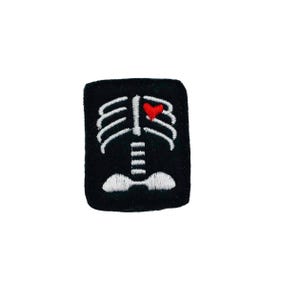 May include: Black embroidered patch with a white skeleton outline and a red heart in the chest area.