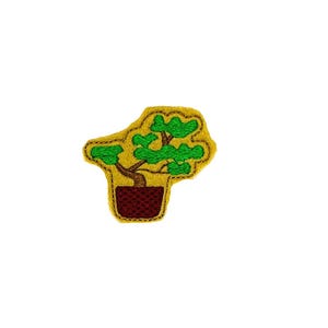 May include: A green and brown felt applique of a bonsai tree in a brown pot. The tree is on a yellow felt background.