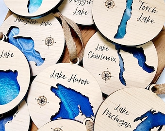 Puede incluir: Un conjunto de adornos de madera con contornos de los Grandes Lagos, cada uno con una brújula y el nombre del lago. Los adornos están pintados en tonos de azul y blanco, que se asemejan al agua.