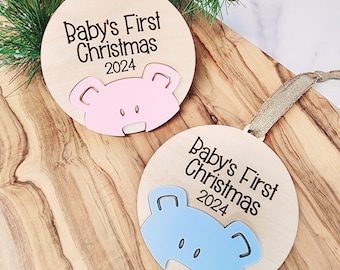 May include: Two wooden Christmas ornaments with a bear head cutout. The ornament on the left has a pink bear head and says "Baby's First Christmas 2024". The ornament on the right has a blue bear head and says "Baby's First Christmas 2024".