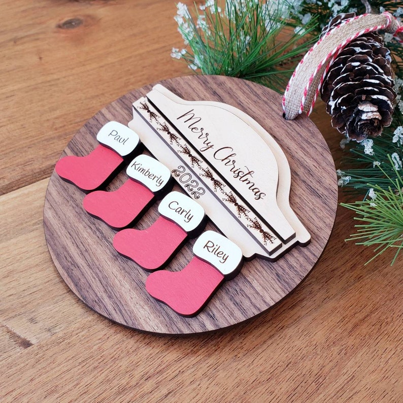 May include: A wooden Christmas ornament with a red truck design. The truck has a white roof with the words "Merry Christmas" and the year "2022" written on it. There are four red stockings hanging from the truck, each with a name written on it: Paul, Kimberly, Carly, and Riley.