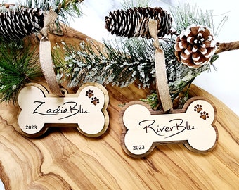 May include: Two wooden dog bone ornaments with the names "Zadie Blu" and "River Blu" engraved on them. The ornaments are decorated with paw prints and the year "2023".