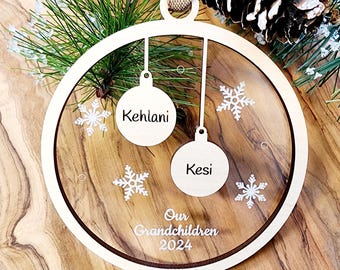 May include: A wooden Christmas ornament with a clear circle in the center. The circle has two small ornaments with the names "Kehlani" and "Kesi" written on them. The circle also has snowflakes and the text "Our Grandchildren 2024".
