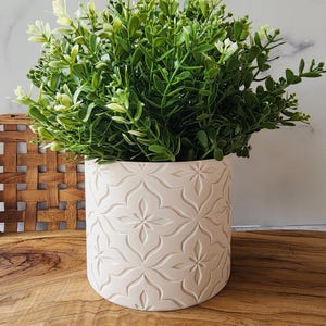 May include: A cylindrical white planter featuring a repeating embossed floral pattern on its exterior. The planter holds a dense, bushy artificial plant with small, vibrant green leaves and light-colored tips, creating a full, rounded appearance.