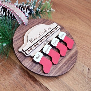 May include: A wooden Christmas ornament with a car silhouette and the text "Merry Christmas 2022".  Four stockings are attached to the bottom with the names "Paul", "Kimberly", "Carly", and "Riley" written on them.